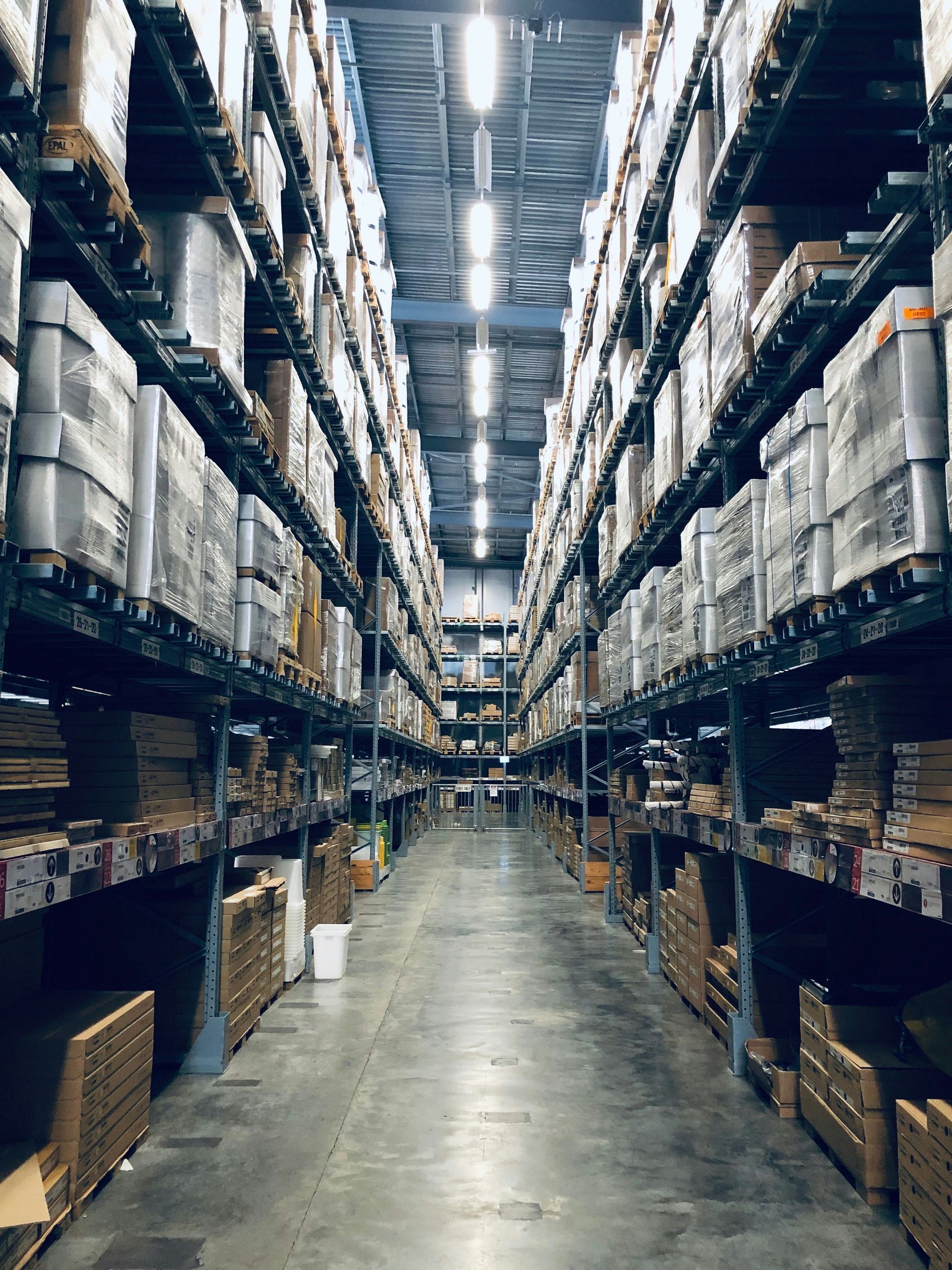 Tall warehouse aisle lined with stacked boxes and shelving under bright overhead lights