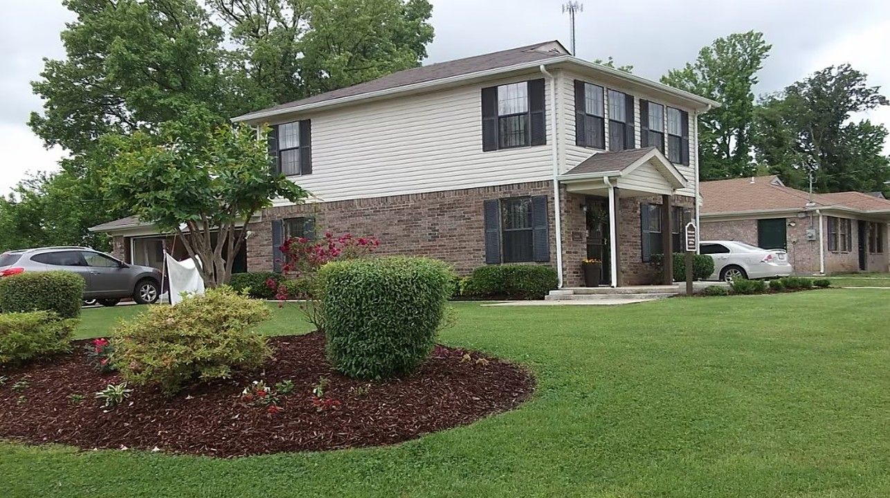 Two-story duplex with beige siding and brick facade. Green lawn and landscaping in front. Cars parked nearby.