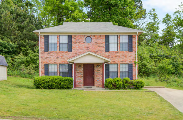 Two-story brick house with black shutters, surrounded by greenery and a lawn.