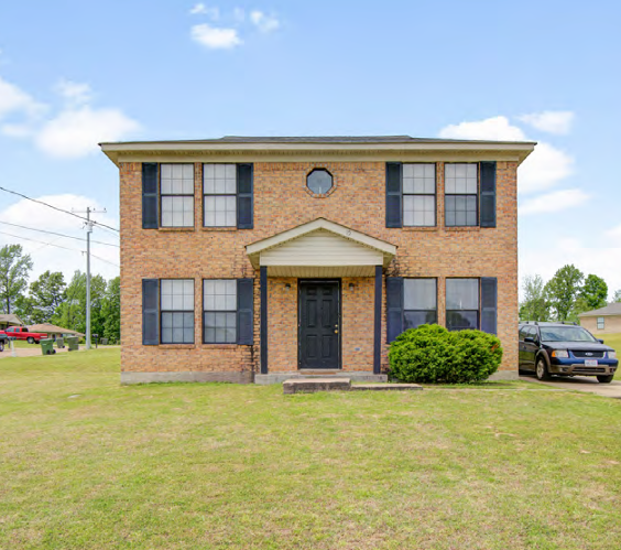 Two-story brick home with black shutters, door, and a car parked nearby on a sunny day.