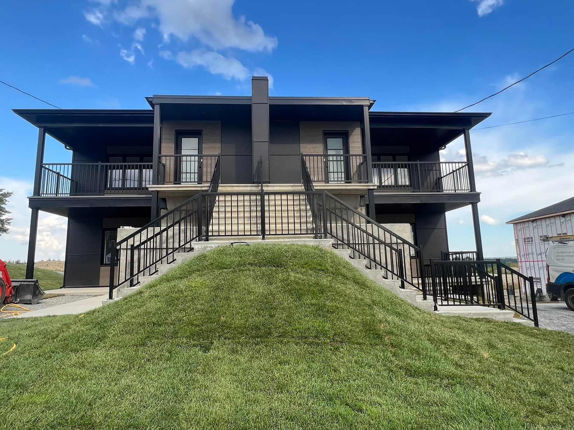 Two-story brown building with balconies, reached by exterior staircases over a grassy mound, blue sky in background.