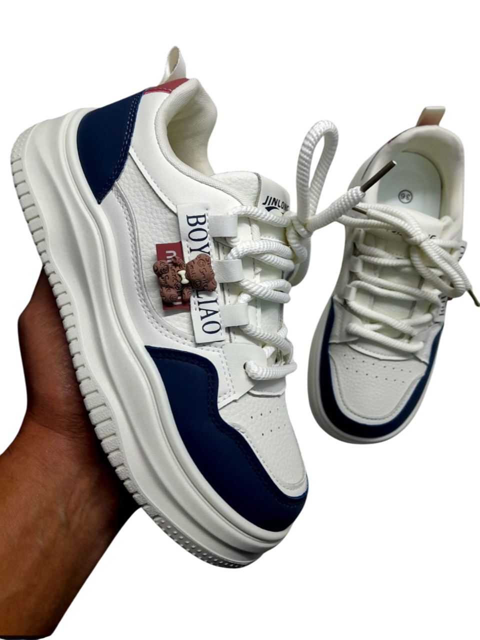Zapatillas blancas y azul marino con suela gruesa blanca, sujetas con una mano. Texto "BOY LAO" en el lateral.