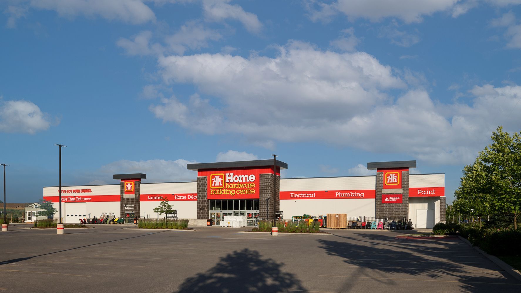 Shopping center under a blue sky. Buildings are white, gray, and brown, with landscaping and parked cars visible.
