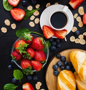 Een bovenaanzicht van een ontbijt met aardbeien, bosbessen, muesli, koffie en brood op een zwarte achtergrond.