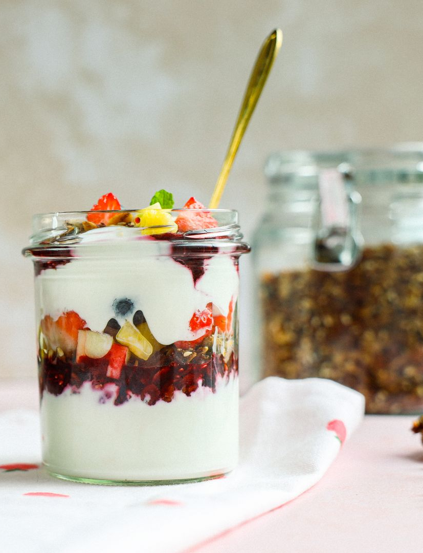 Een glazen pot gevuld met laagjes yoghurt, bessencompote en vers fruit, met daarachter een gouden lepel en een pot granola.