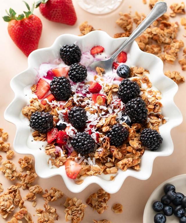 Een witte, gegolfde kom gevuld met yoghurt, granola, bramen en aardbeien, met een lepel en extra fruit.