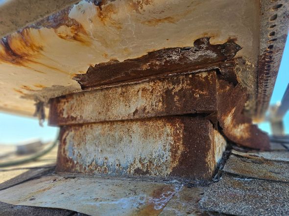 A severely rusted metal roof vent flashing on a shingled roof under a clear blue sky.
