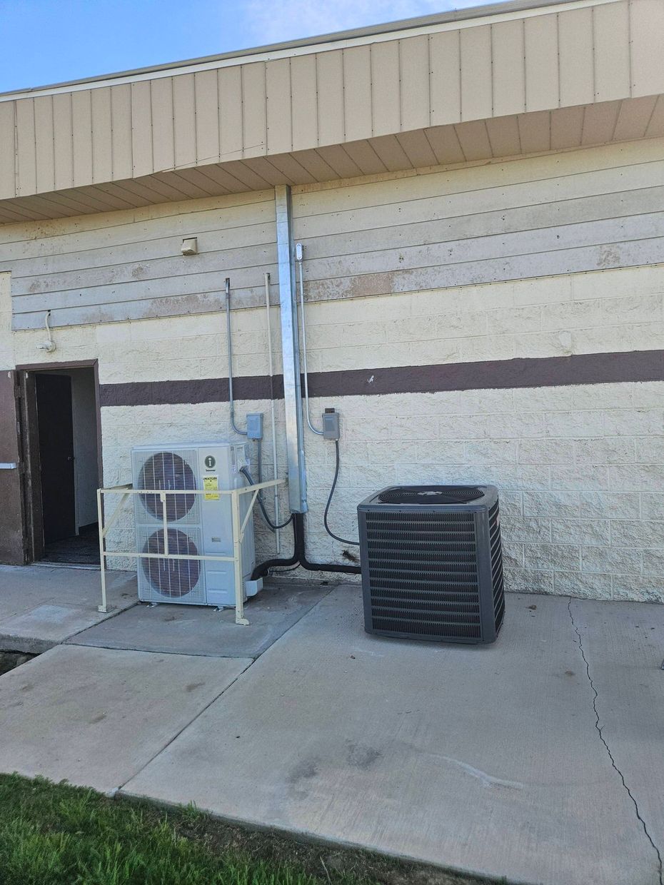 Two HVAC units sit on a concrete pad outside a building wall with a vertical metal duct and an open doorway.