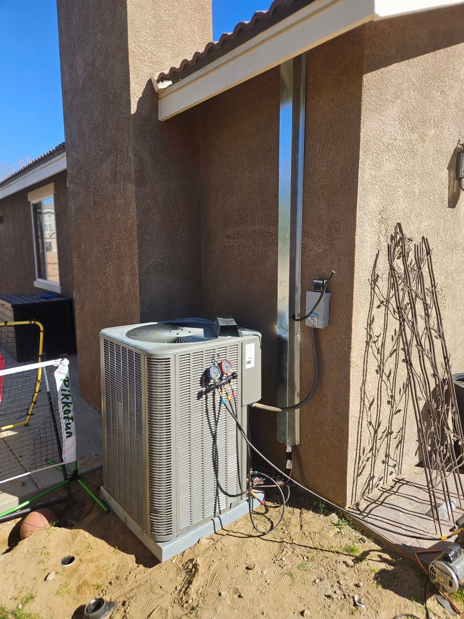 An outdoor air conditioning unit sits against the beige stucco wall of a house with a metal conduit running up the side.