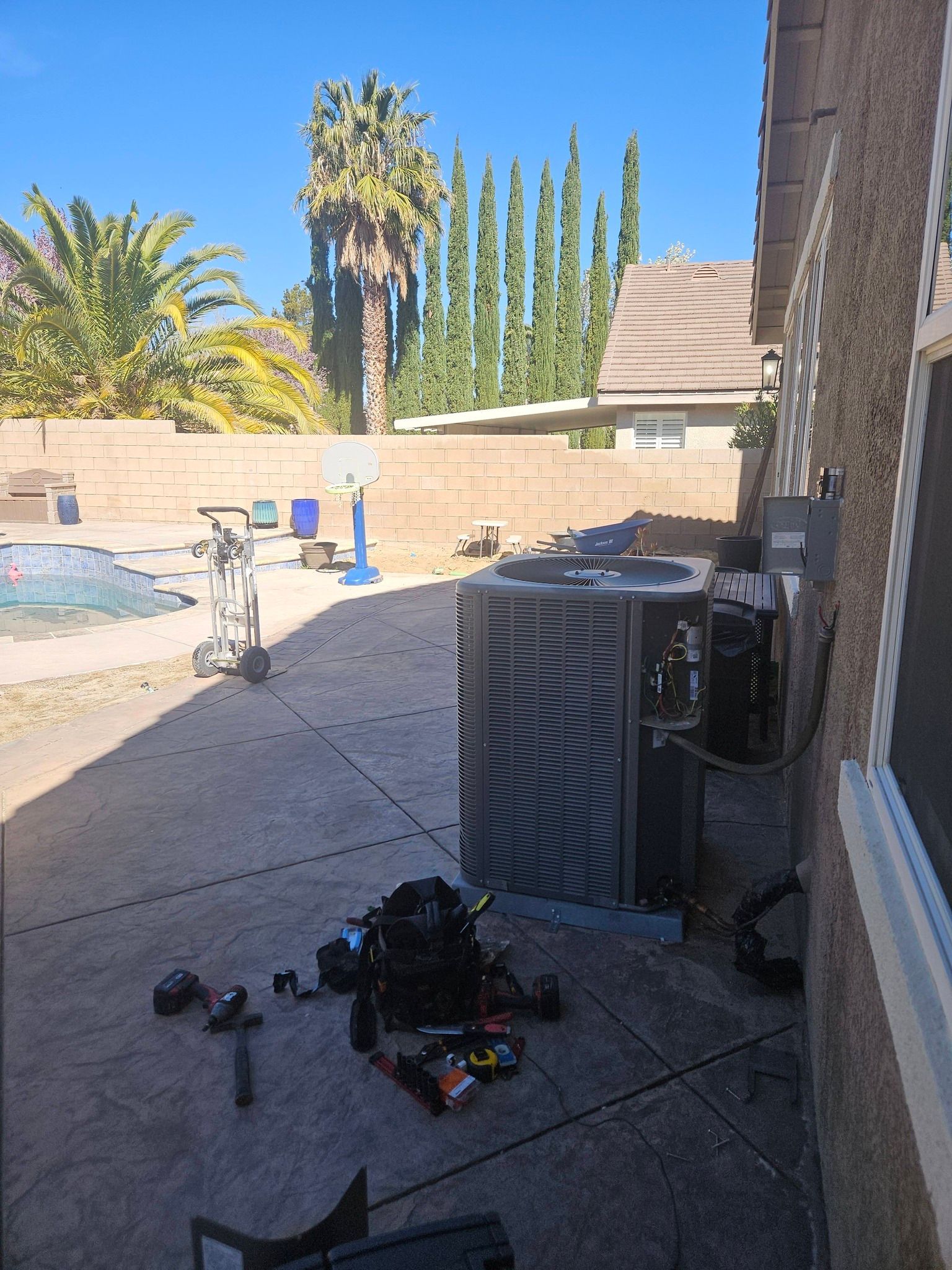 An HVAC technician's tool bag and tools scattered on a patio next to an outdoor air conditioning unit near a pool.