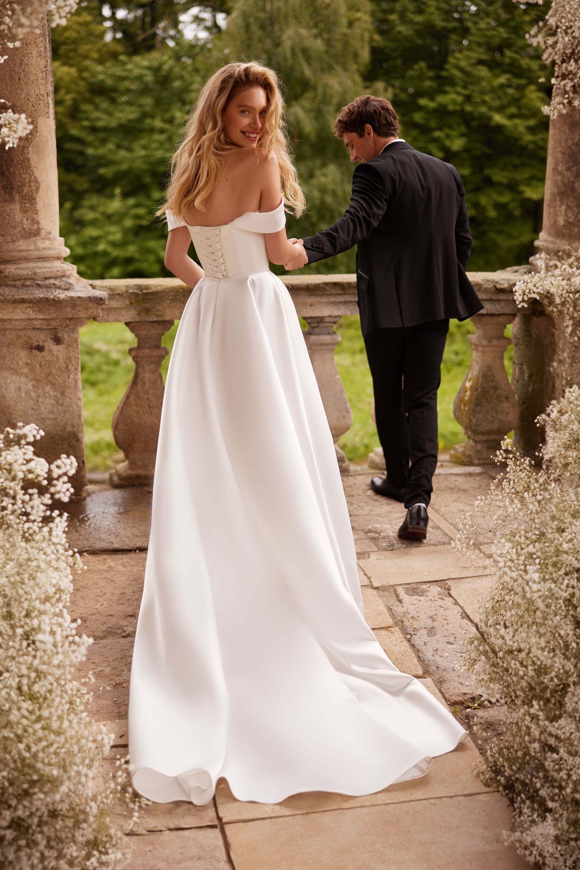 Bride in white dress on a stone path, looking back at groom in black suit, holding hands, outdoor setting.