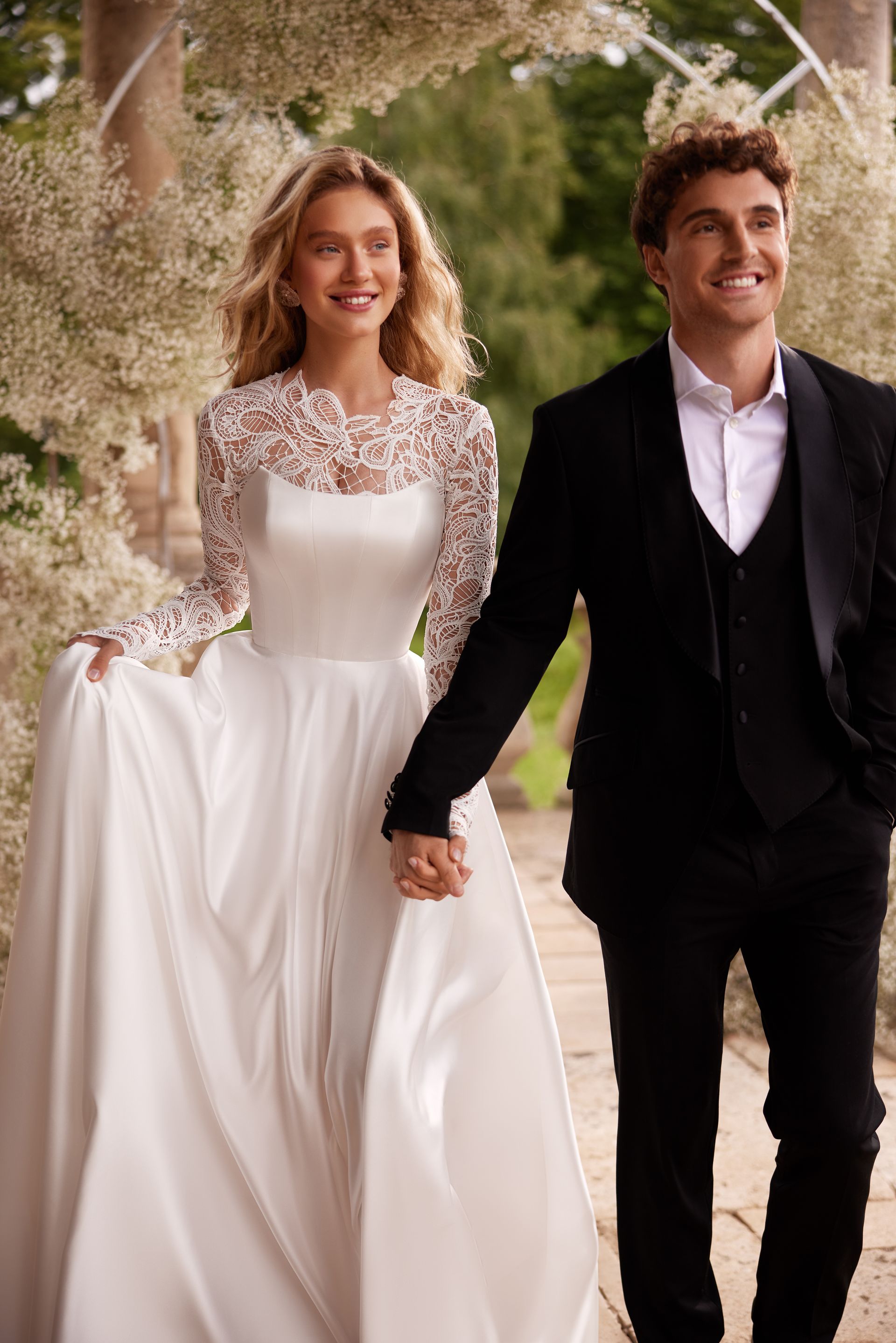 Bride in white dress holds hands with groom in black suit, smiling, walking outdoors.