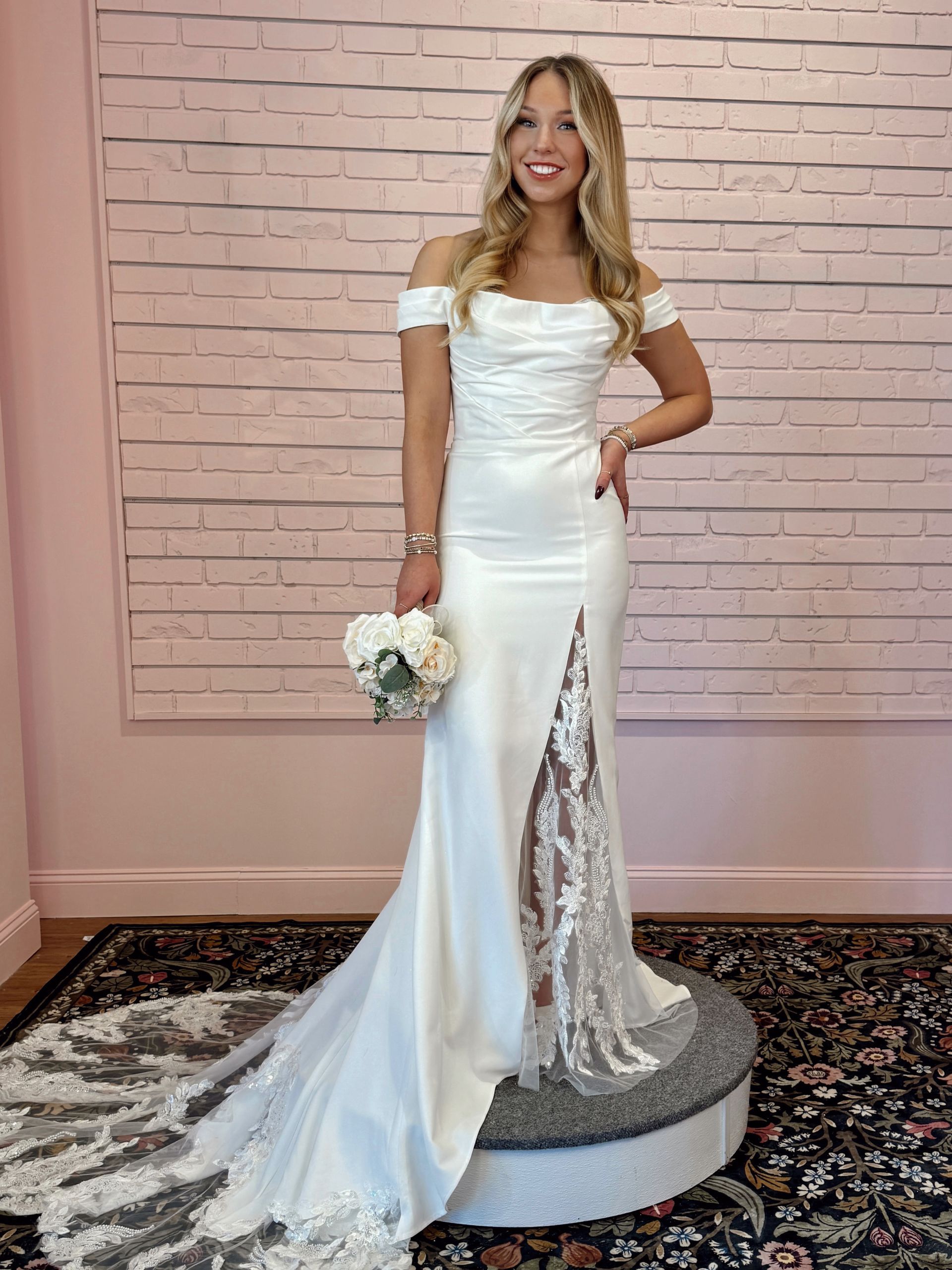 Woman in white off-the-shoulder wedding dress with a thigh-high slit, holding flowers, smiling.