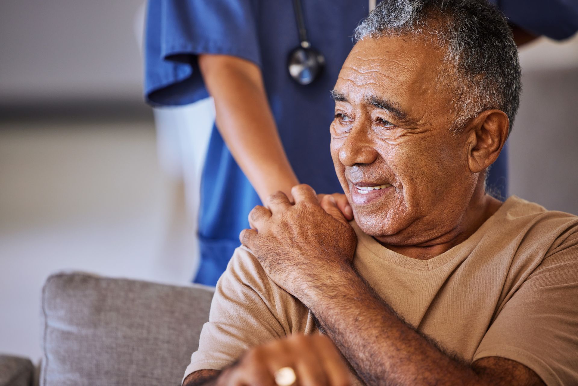 Smiling man seated on a couch, clasping his hands, with a caregiver standing behind him in blue scrubs