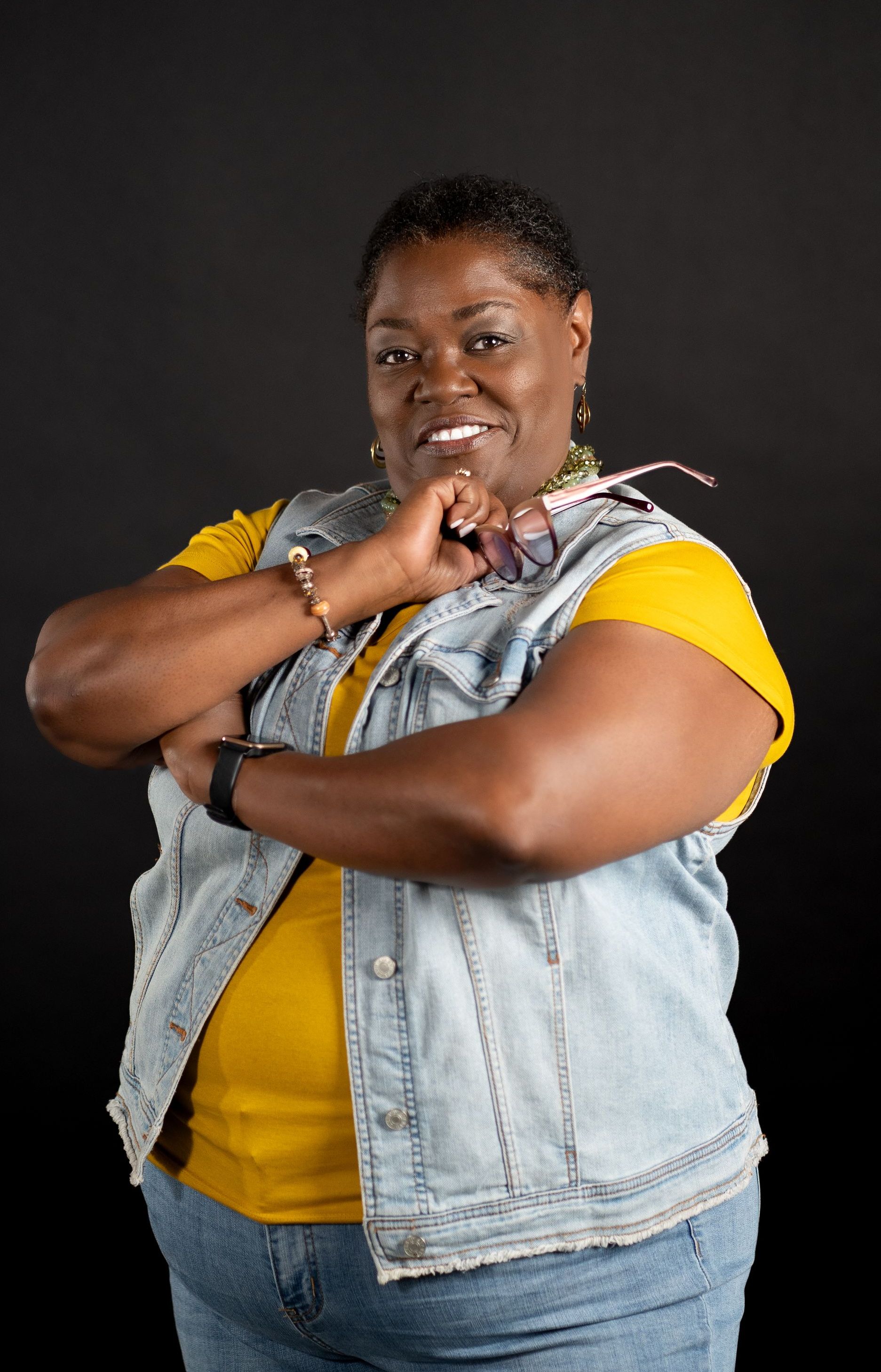 Angela Bailey Page standing holding her glasses on her shoulder with a friendly smile. 