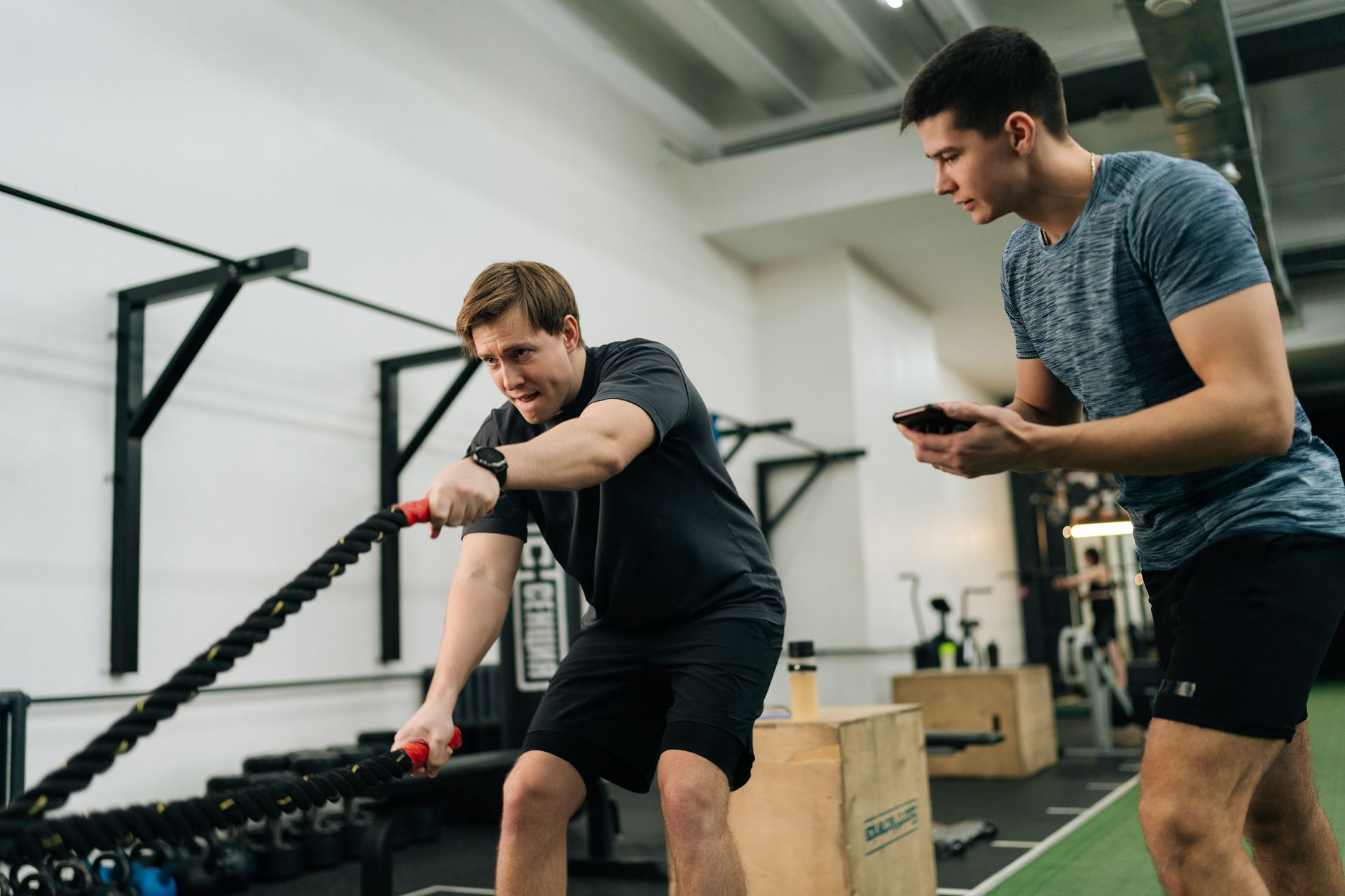 Man exercising with battle ropes, another man holding phone. Gym setting, white walls, black equipment.