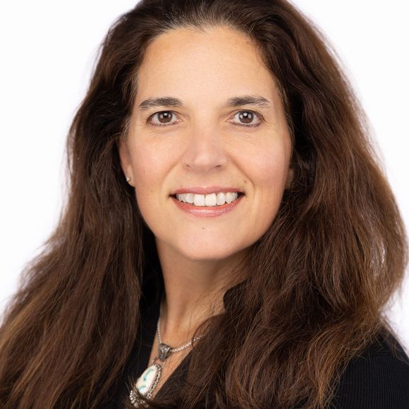 A woman with long brown hair is smiling and wearing a necklace