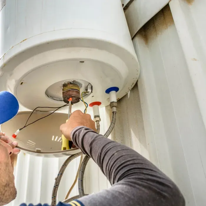 Person repairs a white water heater, working on the exposed underside with a screwdriver.