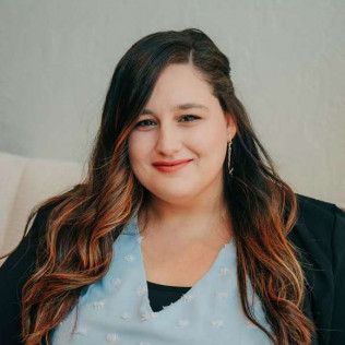 Woman with long brown hair smiling, wearing a blue shirt and black blazer.