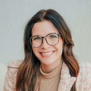 Woman with glasses smiles; wearing a pink jacket and tan turtleneck.