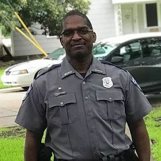 Man in gray police uniform stands outdoors, light skin, glasses, police badge.