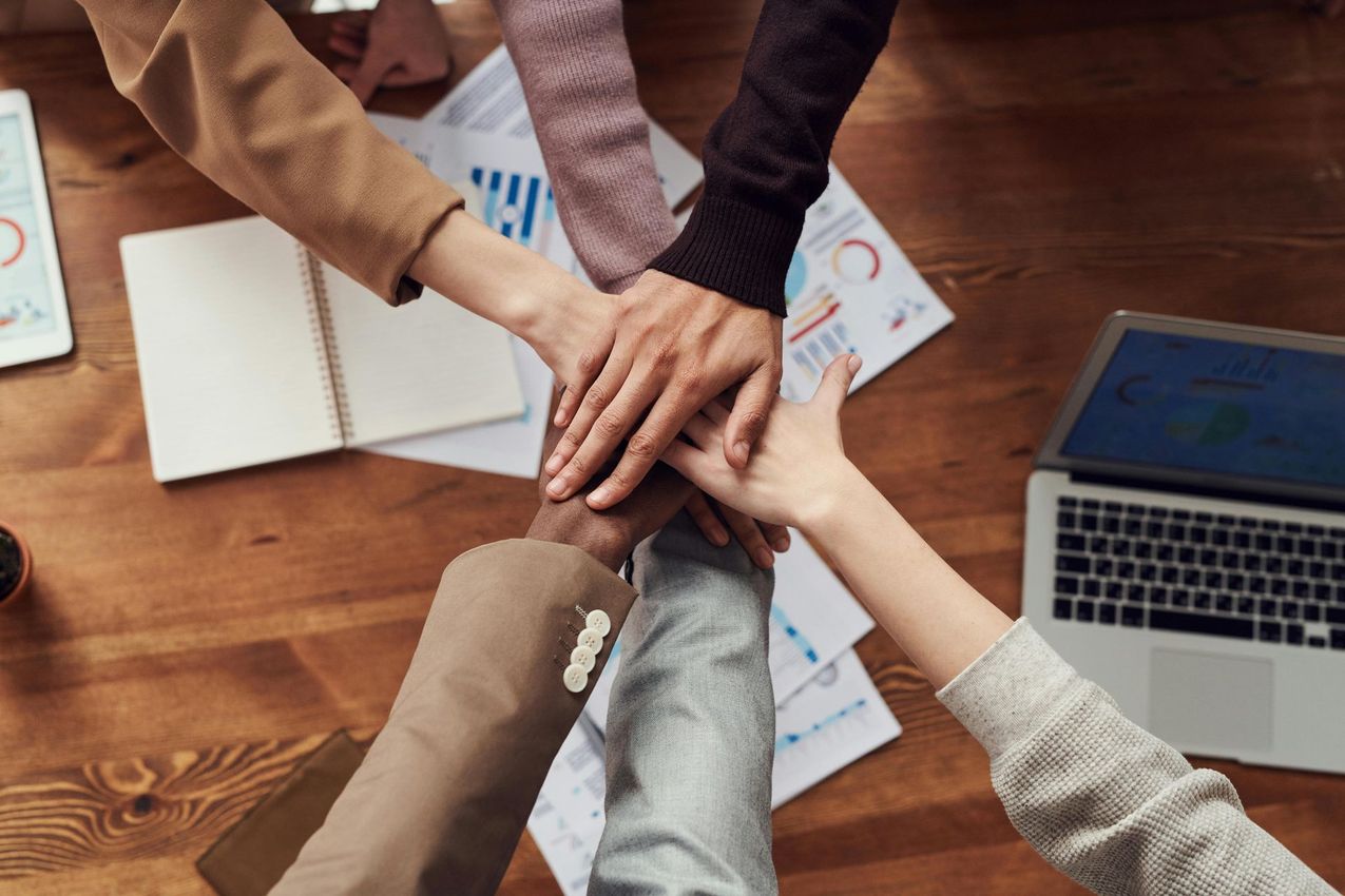 Hands stacked together on a wooden table, above documents and a laptop, symbolizing teamwork.