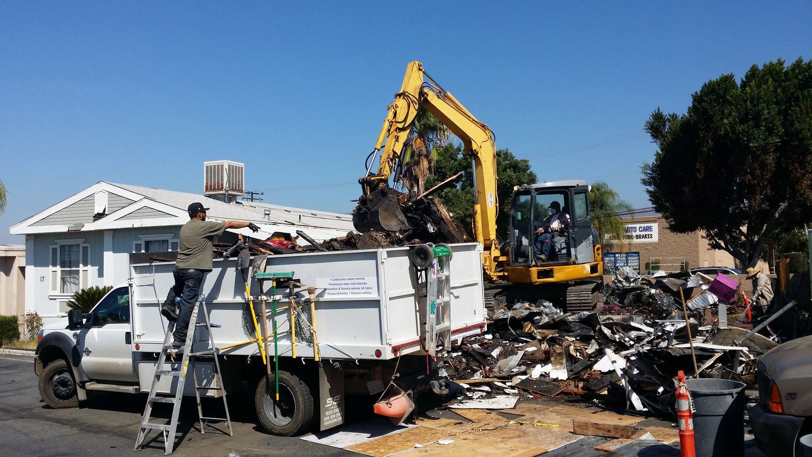 A construction worker loads debris into a white dump truck using a yellow excavator in a residential neighborhood.