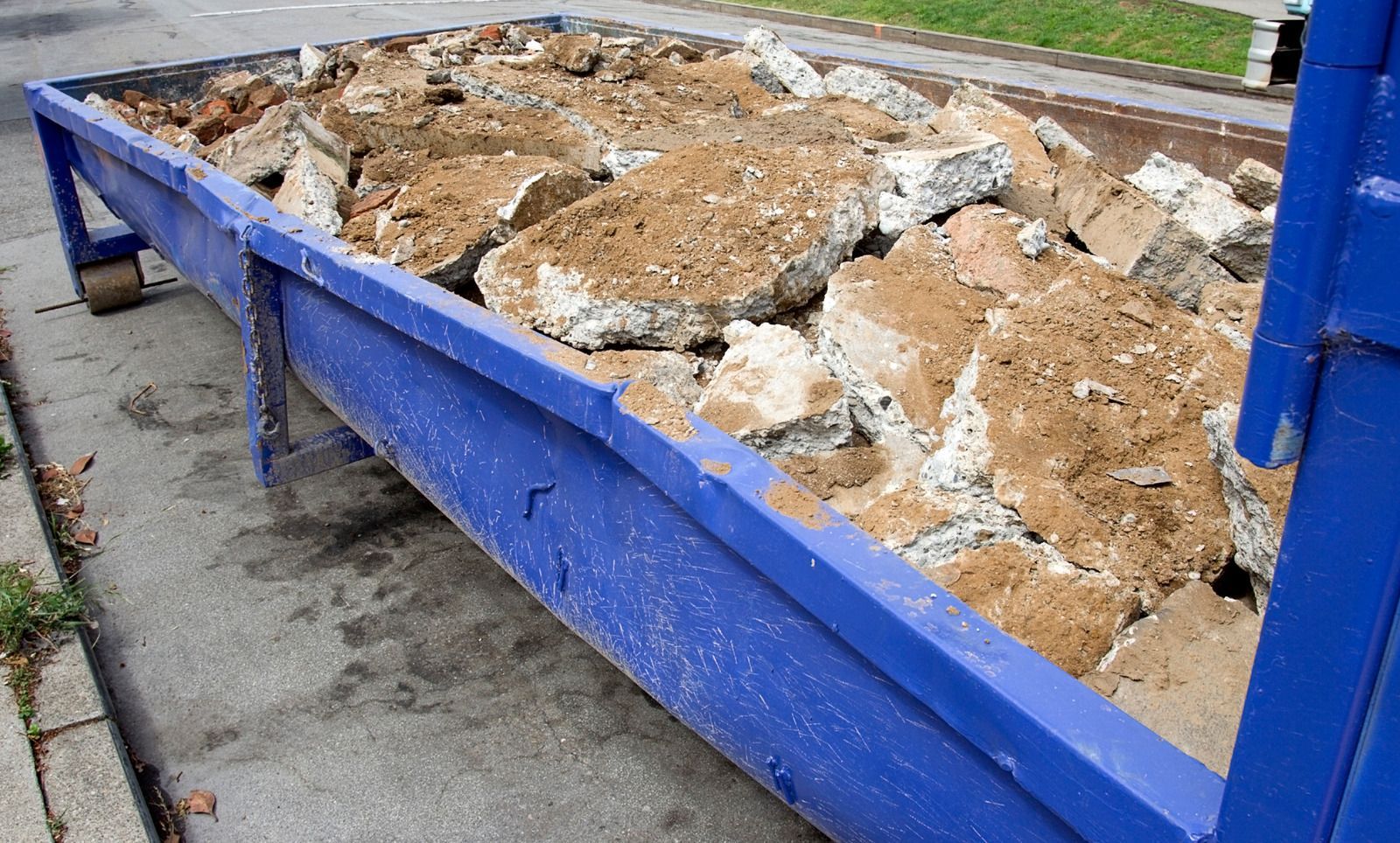 A blue dumpster filled with large pieces of broken concrete and dirt, parked on a paved street.