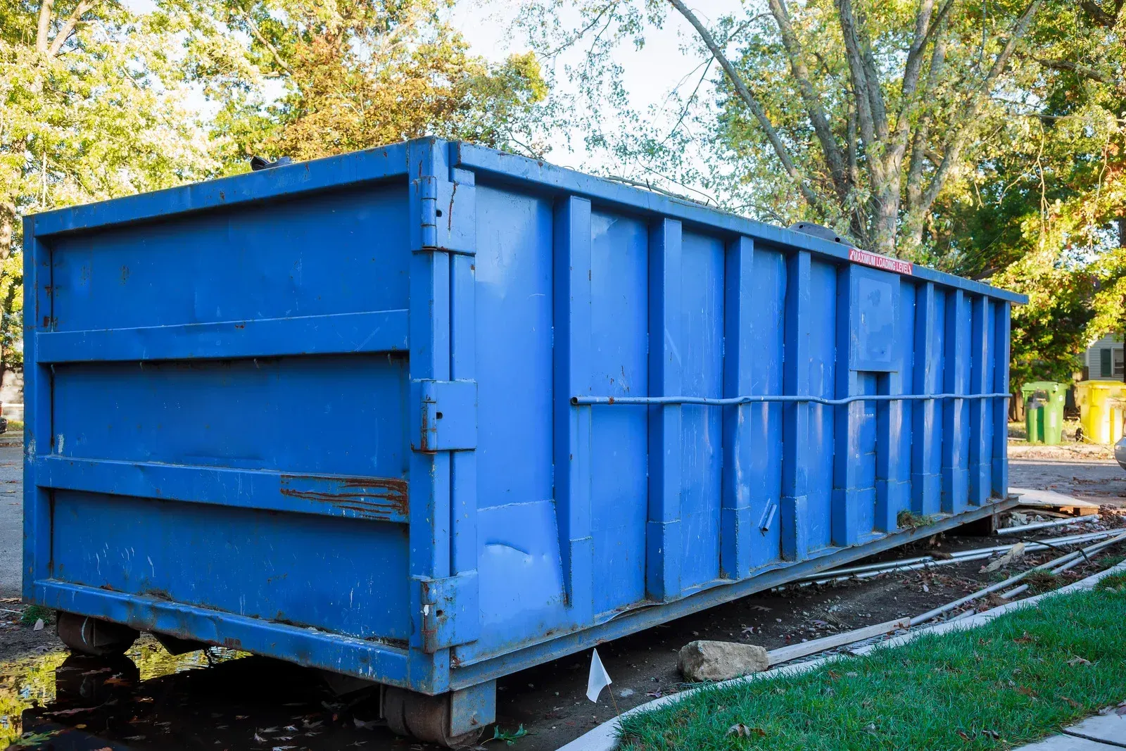 A large blue industrial dumpster sits on a grassy lawn next to a sidewalk with trees in the background.