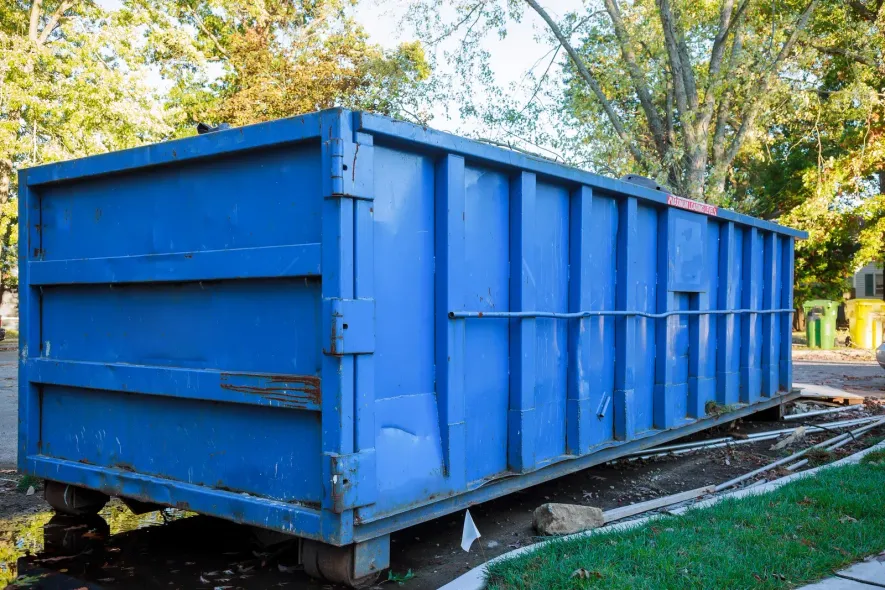 A large blue industrial dumpster sits on a grassy lawn next to a sidewalk with trees in the background.