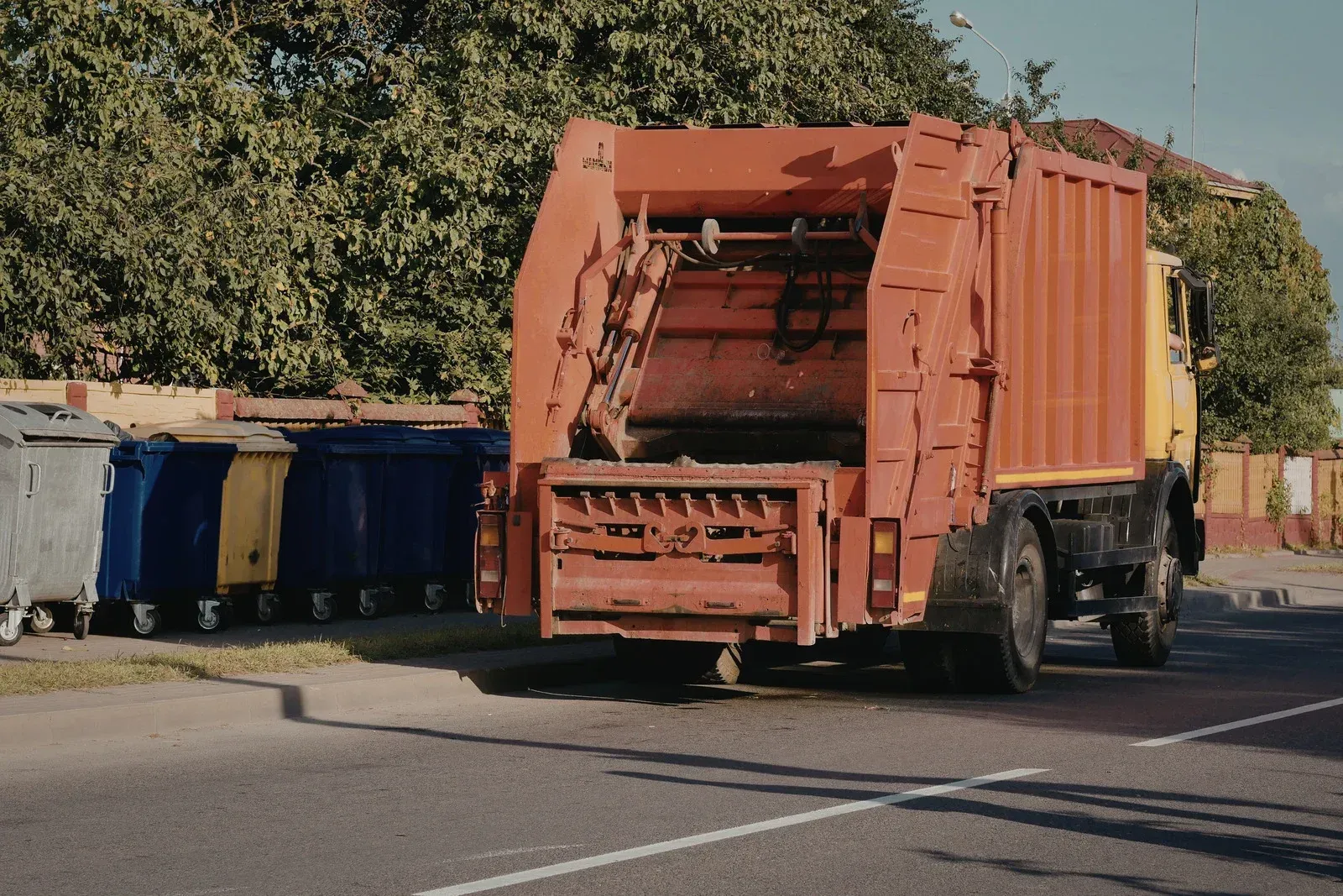A large orange garbage truck parked on a road next to several multi-colored dumpsters on a sunny day.