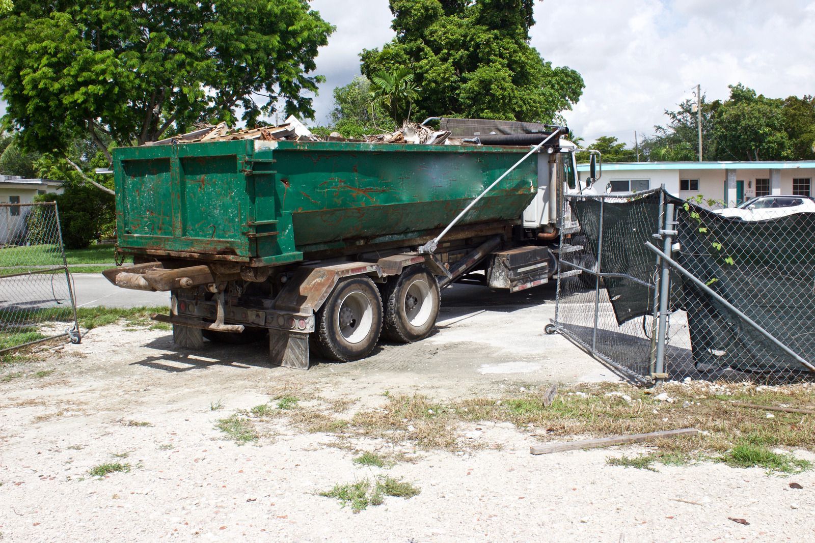 A green roll-off dumpster truck parked on a gravel lot near a black construction fence and residential buildings.