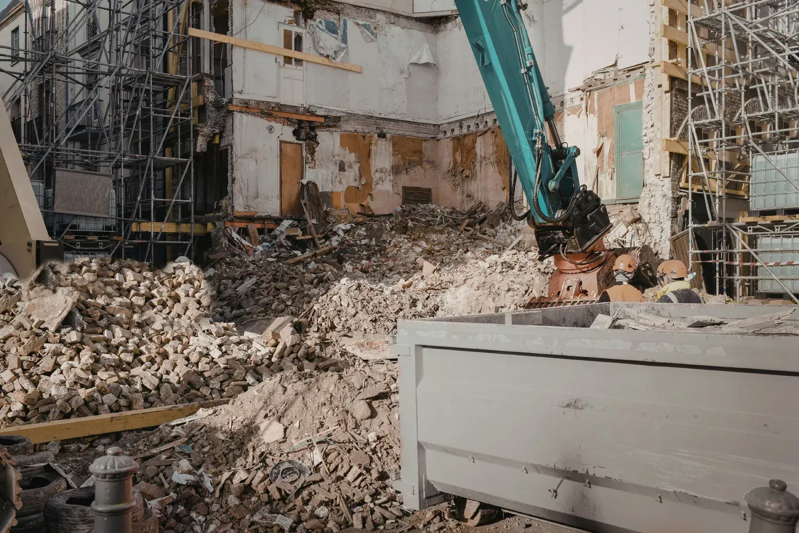 A blue hydraulic excavator demolishes a damaged building, creating a large pile of rubble in a construction site.