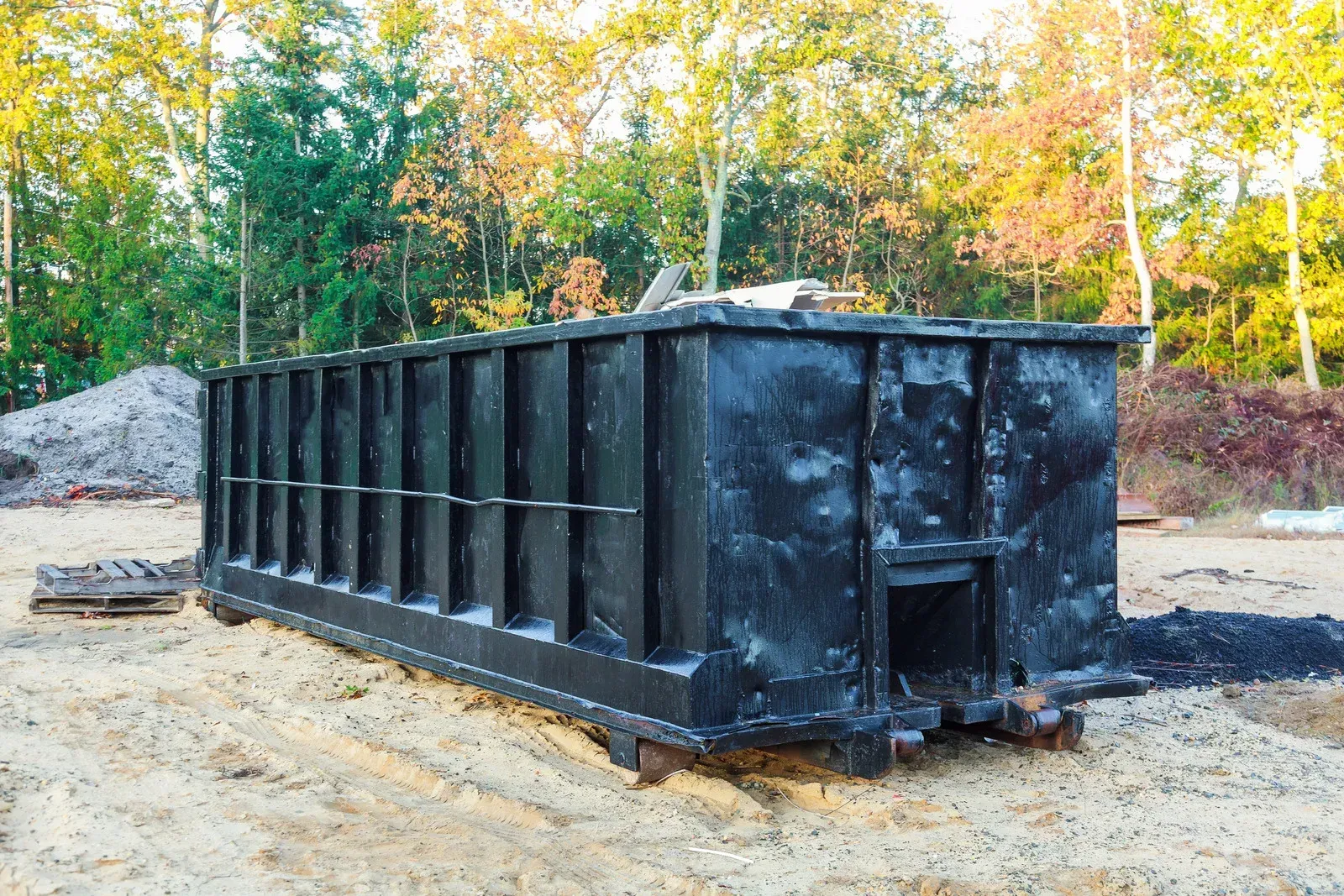 A large, rectangular black metal dumpster sits on a sandy construction site in front of a line of trees.