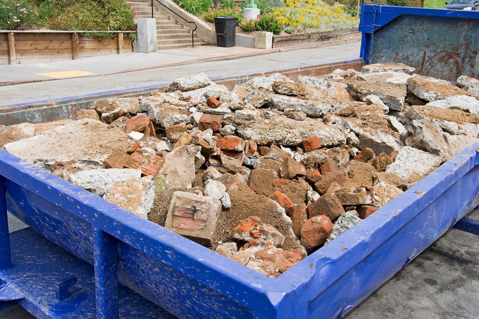 A large blue dumpster filled with concrete rubble, bricks, and construction debris outdoors on a paved surface.