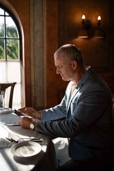 Man in light blue blazer, seated at a restaurant table, looking at his phone. Sunlight streams in from a window.