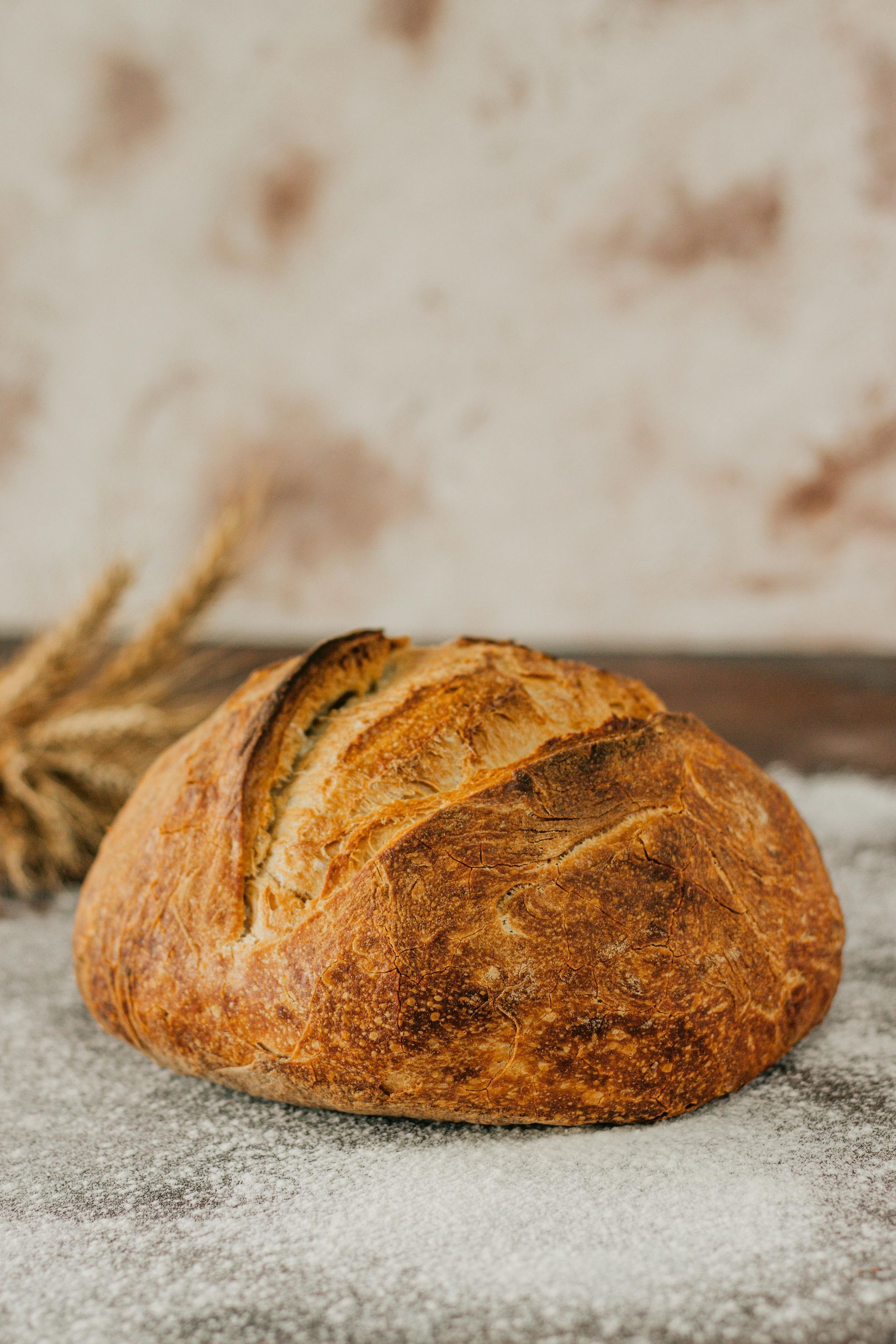 Round loaf of golden-brown artisan bread on a floured wooden surface, with wheat stalks in the background.