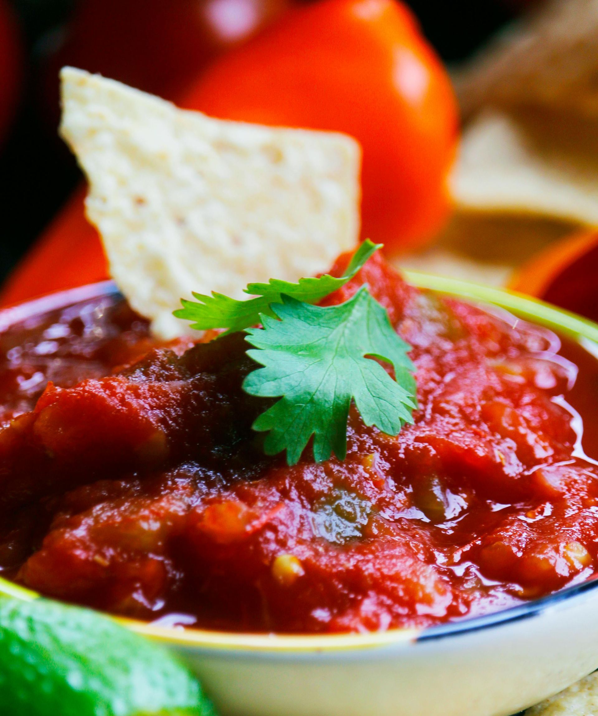 Bowl of red salsa with a tortilla chip and cilantro, surrounded by tomatoes and lime.