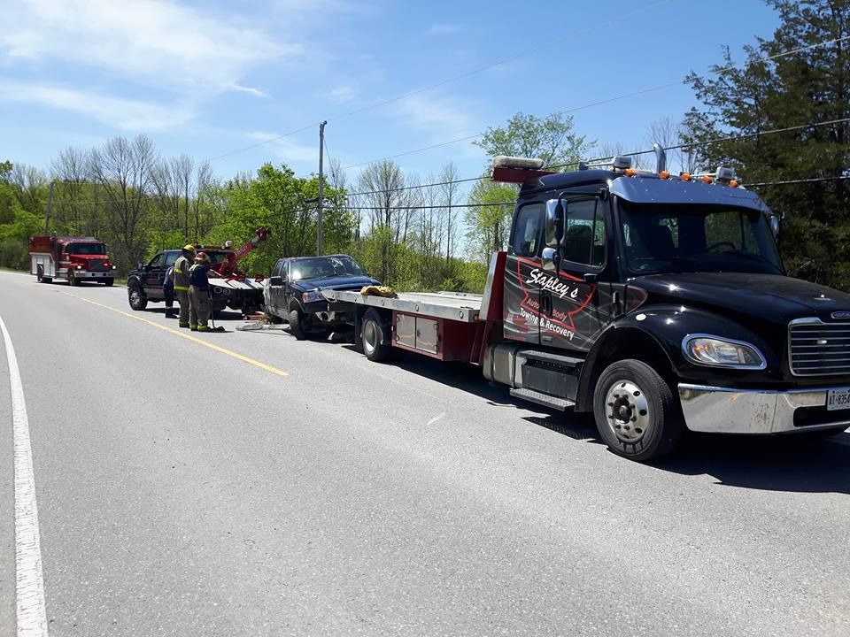 Tow truck hauling two cars on a road. Emergency personnel and fire truck present. Sunny day.