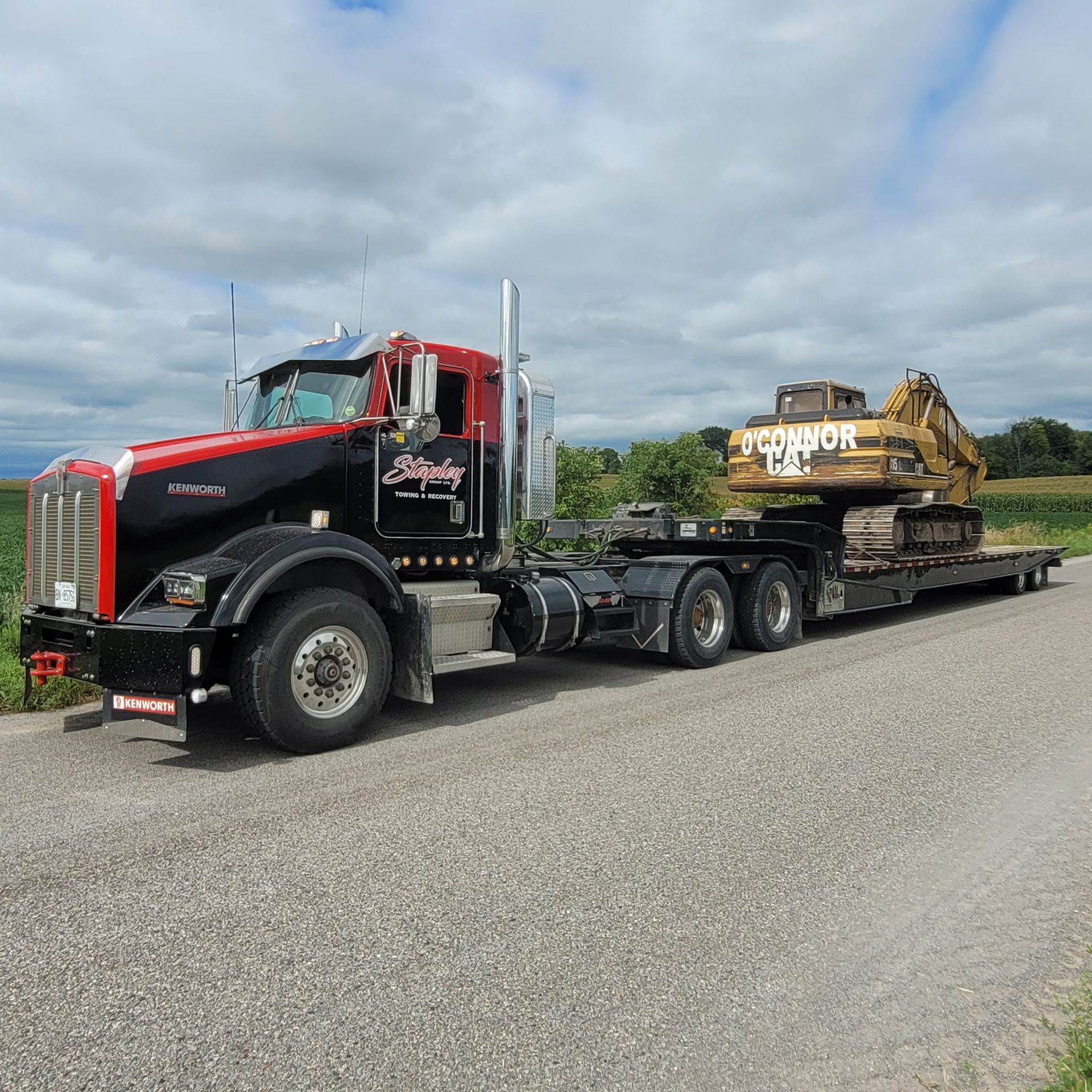 Black and red semi-truck with excavator on a flatbed trailer, parked on a gravel road.