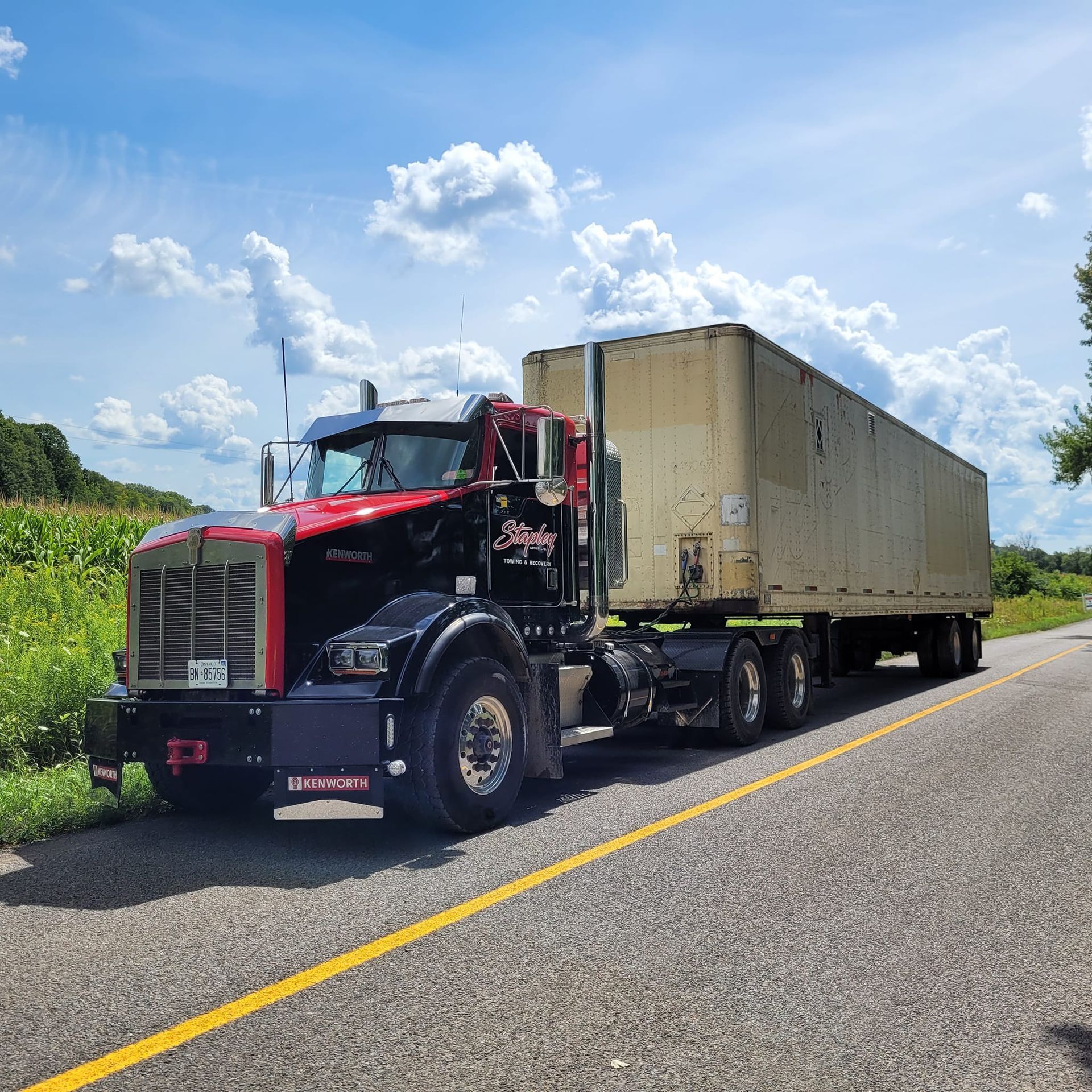 Semi-truck with red and black cab and beige trailer on a road. Bright, sunny day.