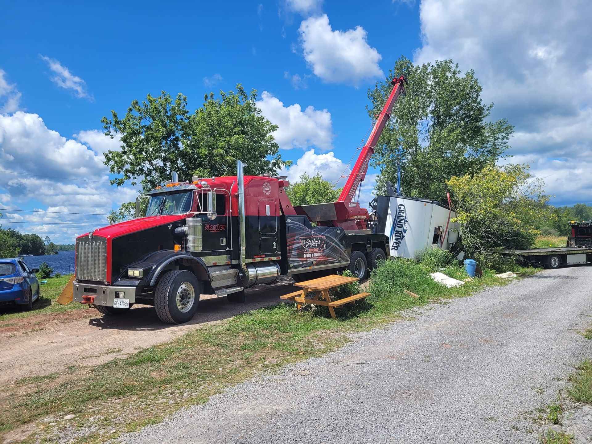 Tow truck lifts a white container near a waterfront, under a blue sky with trees.
