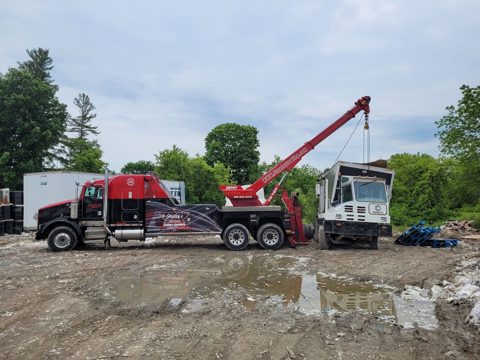 Red tow truck lifting a white industrial vehicle, outdoors on a muddy lot, under a cloudy sky.