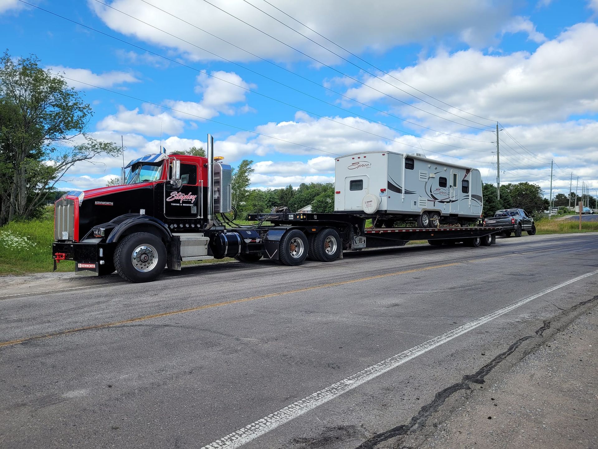 A black semi-truck hauling a white RV on a flatbed trailer on a road under a blue sky.