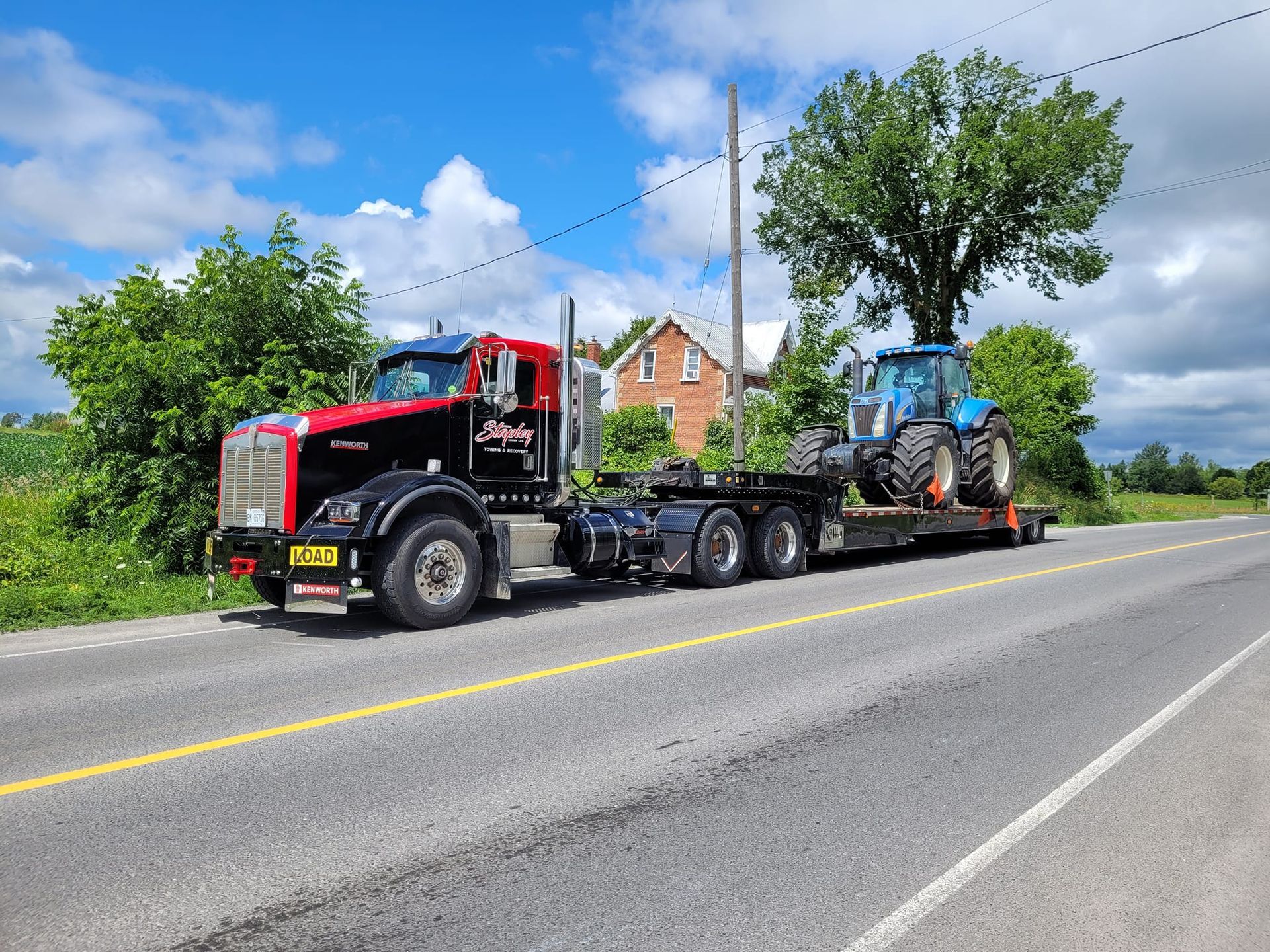 Black and red semi-truck hauling a blue tractor on a trailer along a rural road, under a cloudy sky.