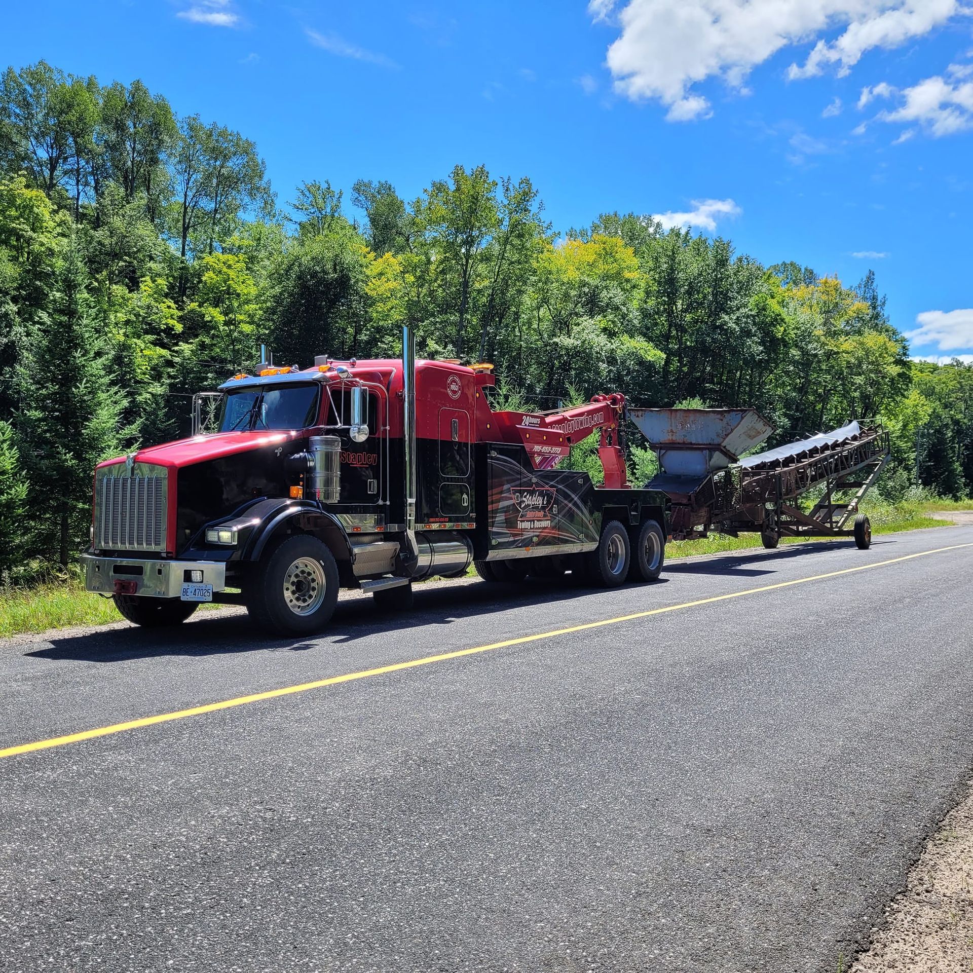 Red and black truck towing a conveyor belt on a paved road with trees and blue sky.