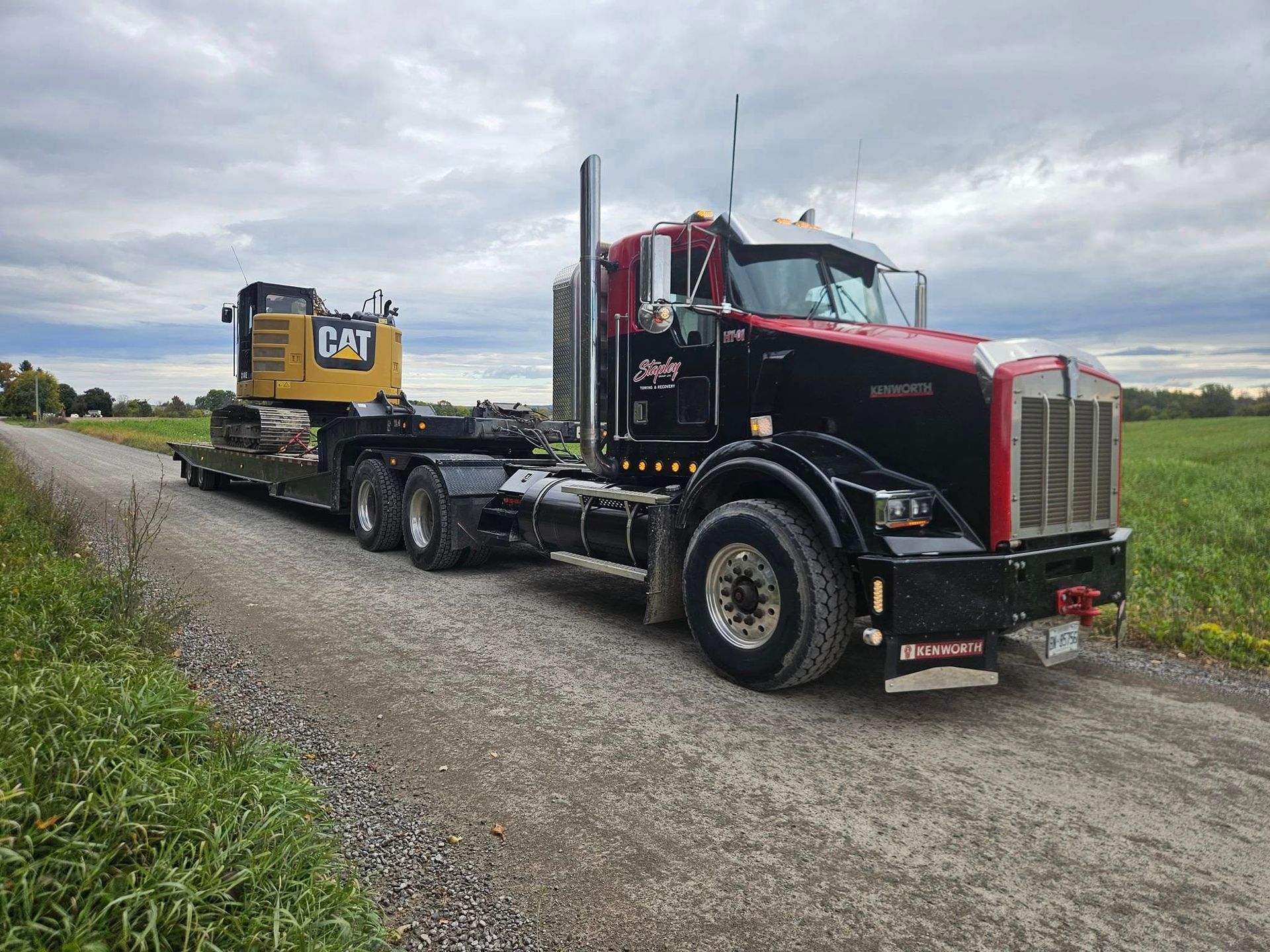 Black and red semi-truck with a yellow excavator on a flatbed trailer on a gravel road in a field.