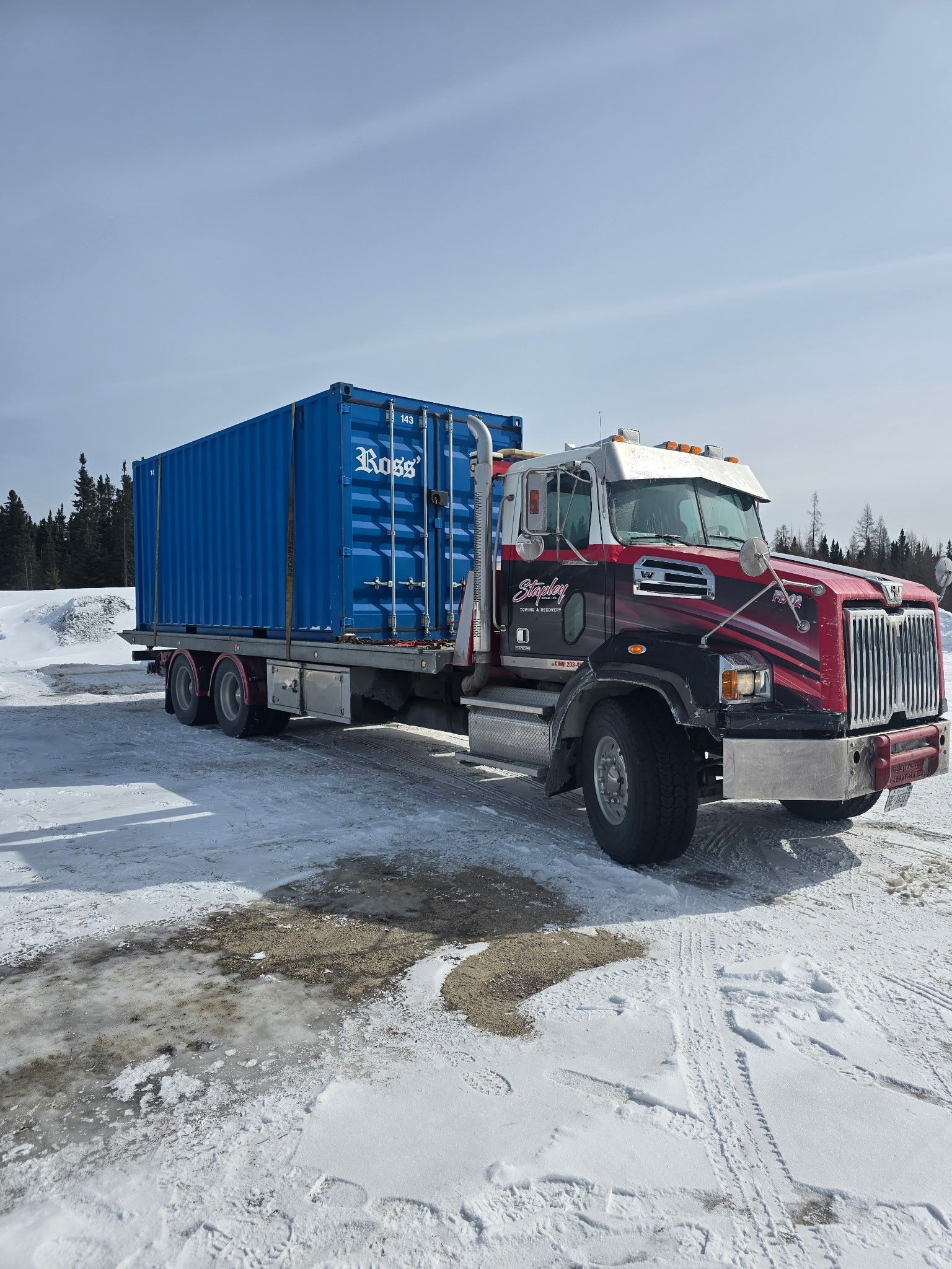 Blue container on a red and black semi-truck parked in a snowy area on a sunny day.