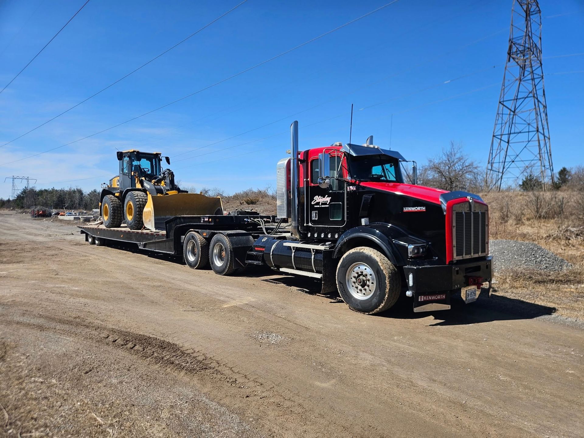 Black and red semi-truck with a front-end loader on a flatbed trailer on a dirt road under a blue sky.