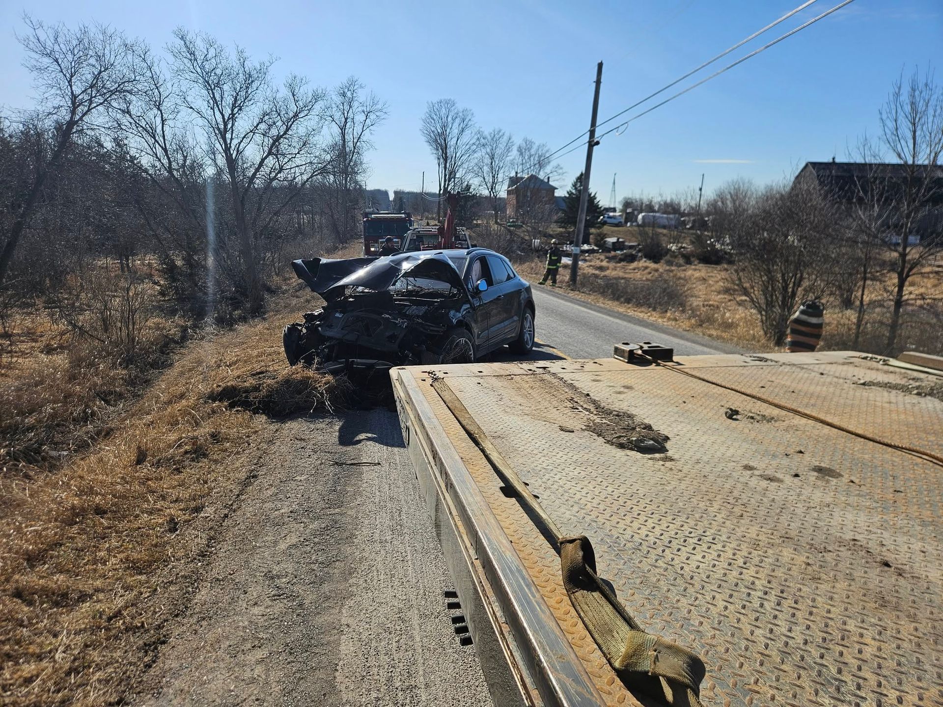 Damaged black car on tow truck next to a rural road, sunny day.